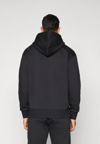 Homem vestindo um hoodie preto liso e calças pretas combinando, de costas para a câmara contra um fundo cinza-claro.