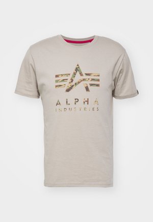 Beige cotton t-shirt featuring a camo-patterned logo of "ALPHA INDUSTRIES" with five horizontal stripes above, short sleeves, and round neckline.