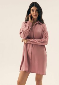 Różowa satynowa sukienka na guziki, z klasycznym kołnierzykiem, długimi rękawami i luźnym krojem z bocznymi rozcięciami dla swobody ruchu.