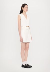 Mujer vestida con un chaleco blanco sin mangas abotonado y una falda plisada, con mocasines negros y calcetines blancos, de pie contra un fondo blanco liso.