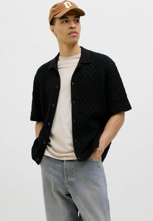 Cardigan noir à manches courtes avec un motif texturé, fermé par des boutons, porté sur un t-shirt clair et assorti à un jean bleu clair et une casquette marron.