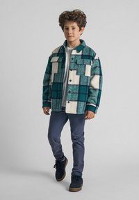 Roheline ja kreemjas ruuduline jakk kahe eesmine taskuga, nööpidega kinni ja kantud valge särk üle, sobitatud tumedate pükste ja sneakeritega.