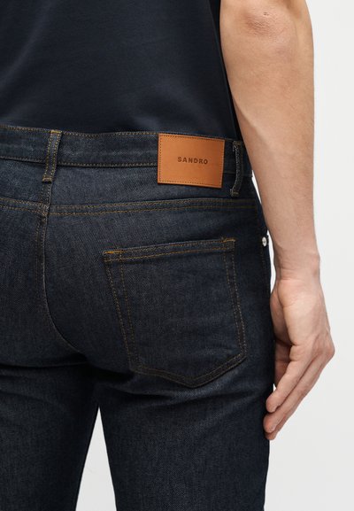 Personne portant un jean bleu foncé avec un patch en cuir marron clair marqué "SANDRO" sur la ceinture, une poche arrière visible et un bras partiellement visible.