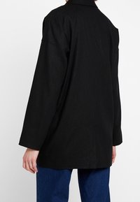 Černá dlouhá košile s rukávy ve formě saka, uvolněného střihu a viditelným šitím. Tkanina vypadá strukturovaně a lehce.