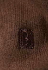 Hnědá pletená tkanina s texturovaným povrchem, která má na svém povrchu tmavohnědé vyšívané logo "B" v čtverci.