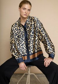 Bluse i leopardmønstret satin med sort og guld farver, knaplukning foran og marineblå detaljer, parret med sorte, løstsiddende bukser.
