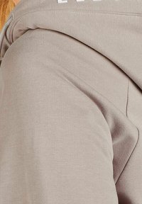 Beżowa bluza z kapturem o miękkiej teksturze, z regulowanym sznurkiem przy kapturze i białym nadrukowanym tekstem na górze. Prosty design bez wyraźnych wzorów.