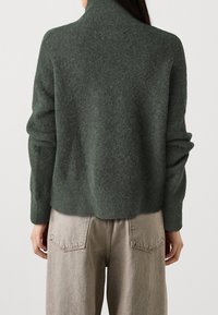 Zielony sweter z mieszanki wełny o wysokim dekolcie i luźnym kroju, z prążkowanymi mankietami i miękkim, teksturowanym wykończeniem. Noszony z jasnobeżowymi dżinsami.