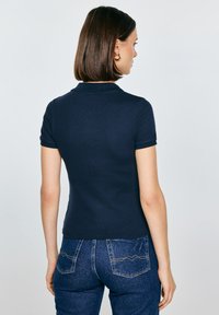 Námořnicky modrá polo košile s krátkým rukávem, přiléhavým střihem a žebrovaným límečkem. Je kombinována s džínami s ozdobnými kapsami na zadní straně.