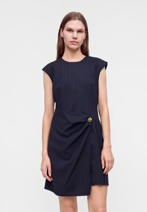 Mujer con un vestido negro sin mangas con detalle de botón dorado y tela fruncida en la cadera izquierda, de pie frente a un fondo liso.