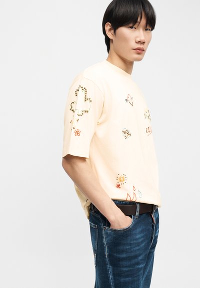 Νεαρό άτομο με μαύρα μαλλιά που φοράει μια εκρού φαρδιά μπλούζα με διακοσμητικά και κεντημένα πολύχρωμα σχέδια, σε συνδυασμό με μπλε τζιν παντελόνι.