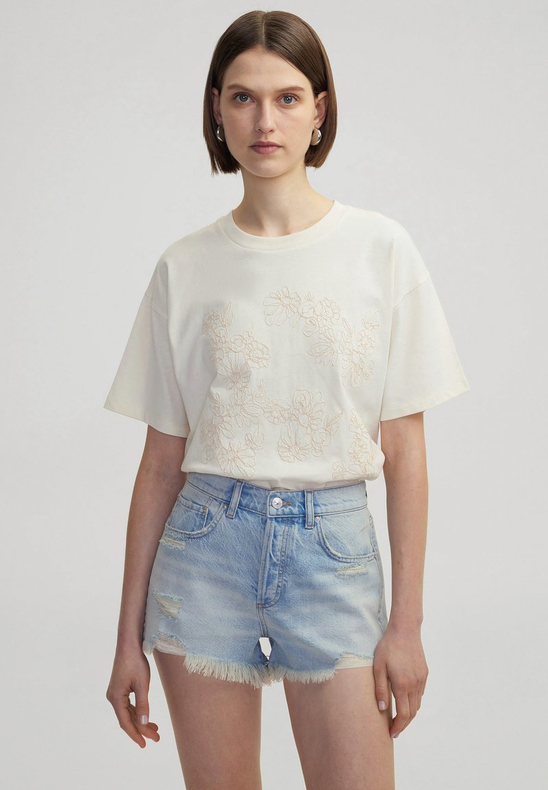 Crème T-shirt met korte mouwen en bloemenborduurwerk aan de voorkant, gecombineerd met lichtblauwe denim shorts met een rafelige zoom en versleten details.