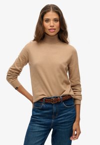 Donna che indossa un maglione beige a collo alto infilato in jeans blu con una cintura intrecciata marrone, in piedi davanti a uno sfondo neutro.