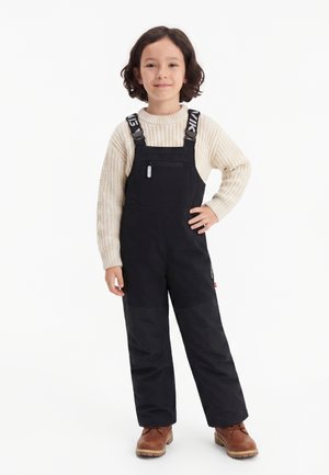 Criança com cabelo castanho escuro de comprimento médio está a sorrir, vestindo um camisola de malha creme, macacão preto com bolso de fecho éclair e botas castanhas.