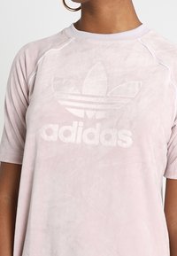 Jasnoróżowa koszulka Adidas z delikatnym, wytłoczonym logo i kwiatowym wzorem. Posiada krótkie rękawy oraz szwy raglanowe, co zapewnia luźny krój.