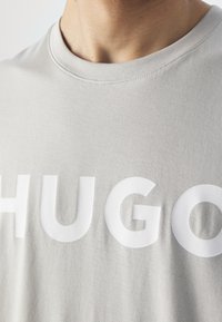 Detailní záběr osoby v světle šedé košili s velkými bílými písmeny "HUGO" vytištěnými na prsou.