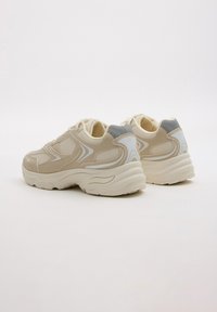 Zapatillas beige con parte superior de malla texturizada, detalles lisos y suelas blancas gruesas. Presenta un talón gris y líneas decorativas sutiles.