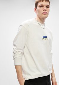 Λευκό μακρυμάνικο μπλουζάκι με στρογγυλή λαιμόκοψη. Διαθέτει μπλε και κίτρινο λογότυπο "HUGO" και "Jujutsu Kaisen" στο στήθος. Μαλακό ύφασμα από βαμβάκι.