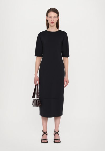 Schwarzes, tailliertes Kleid mit kurzen Ärmeln, glattem Stoff und knielangem Saum, kombiniert mit einer kleinen dunklen Handtasche und schwarzen Riemchensandalen.