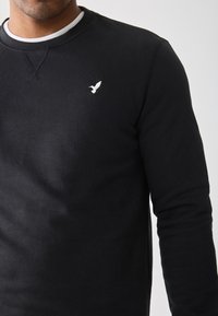 Suéter negro de algodón, con cuello redondo, mangas largas y un pequeño logo de un pájaro blanco en el lado izquierdo del pecho. Textura suave.