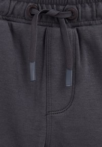 Pantalons de jogging gris foncé avec une taille élastiquée et un cordon de serrage. Présente une texture lisse, bande élastique et un détail de couture discret près des poches.