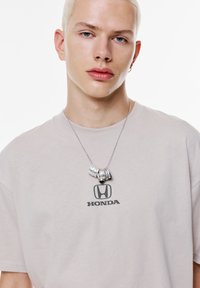 Lys grå t-shirt med sort Honda-logo, sølv halskæde med tre strukturerede, buede metalstykker, på model med kort lyst hår.