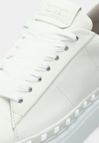 Λευκό δερμάτινο αθλητικό παπούτσι με υφασμάτινα κορδόνια, επίπεδο σχήμα, επενδεδυμένο κολάρο και υφασμένο πάτο με λευκές τρούκς κατά μήκος των πλευρών.