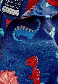 Veste imperméable avec un imprimé de dinosaure bleu sur fond marine, comportant des détails d'accent rouge et des gouttes sur une texture lisse.