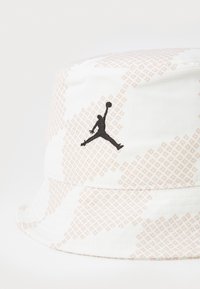 Un chapeau seau blanc avec un motif en rose clair. Un logo Jordan noir est brodé à l'avant. Tissu lisse avec un bord structuré.