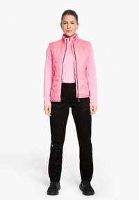 Pink, quilted sleeveless vest over a long-sleeve light pink top, paired with black pants featuring textured panels and black shoes.