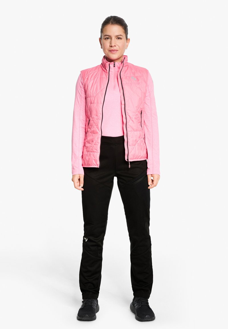 Pink, quilted sleeveless vest over a long-sleeve light pink top, paired with black pants featuring textured panels and black shoes.