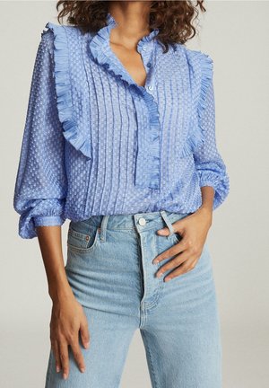 Kobieta ubrana w prześwitującą, jasnoniebieską bluzkę z falbankami i fakturowanymi kropkami, w połączeniu z wysokim stanem jasnoniebieskich dżinsów.