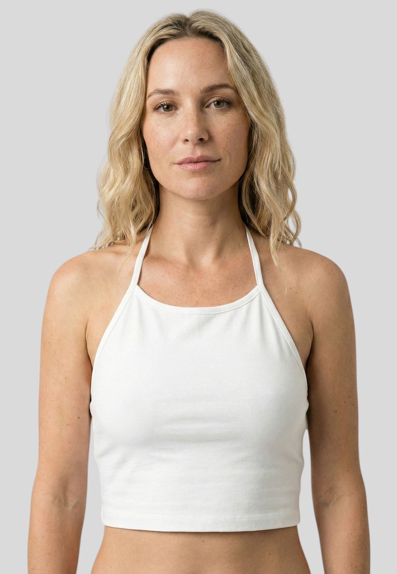 Femme blonde aux cheveux ondulés portant un crop top blanc sans manches, tournée vers l'avant sur un fond gris neutre.