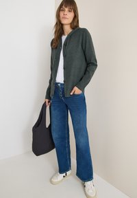 Dunkelgrüner gestrickter Cardigan mit Kapuze und Knöpfen, getragen über einem weißen T-Shirt und kombiniert mit blauen Schlaghosen und weißen Sneakers.