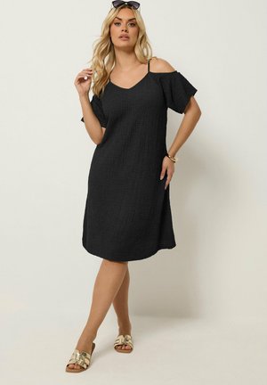 Femme portant une robe midi noire texturée avec épaules dénudées, sandales plates dorées, lunettes de soleil sur la tête, et bracelet doré, posant avec la main près des cheveux.