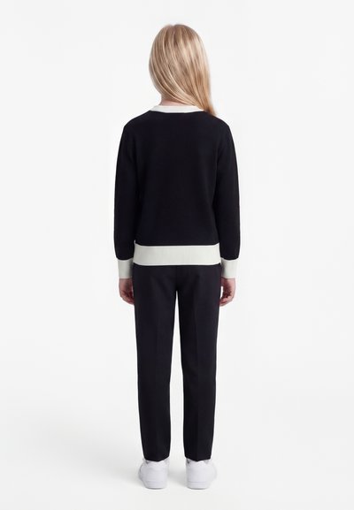 Barn med langt blondt hår står med ryggen mot, iført en svart genser med hvite mansjetter og svarte bukser med hvite joggesko.