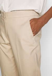 Beżowe, dopasowane spodnie o prostym kroju i gładkiej fakturze. Widoczna jest boczna kieszeń, a tkanina wydaje się lekka i przewiewna.