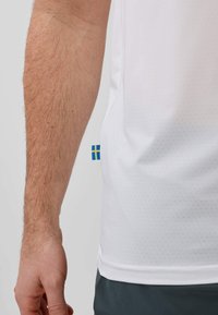 Avant-bras gauche avec des poils visibles à côté d'une chemise blanche perforée montrant une petite étiquette avec le drapeau suédois bleu et jaune sur la couture latérale.