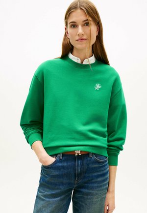 Jeune femme portant un sweat-shirt vert et un jean bleu, la main dans la poche, debout devant un fond blanc uni.