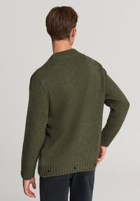 Uomo che indossa un maglione lavorato a maglia color verde oliva con piccoli fori vicino all'orlo, in piedi con le spalle rivolte alla macchina fotografica.