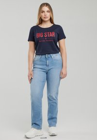 Moteris stovi dėvėdama tamsiai mėlyną marškinėlį su užrašu „BIG STAR“, šviesiai mėlynas džinsus ir baltus sportinius batelius, priešais paprastą pilką foną.