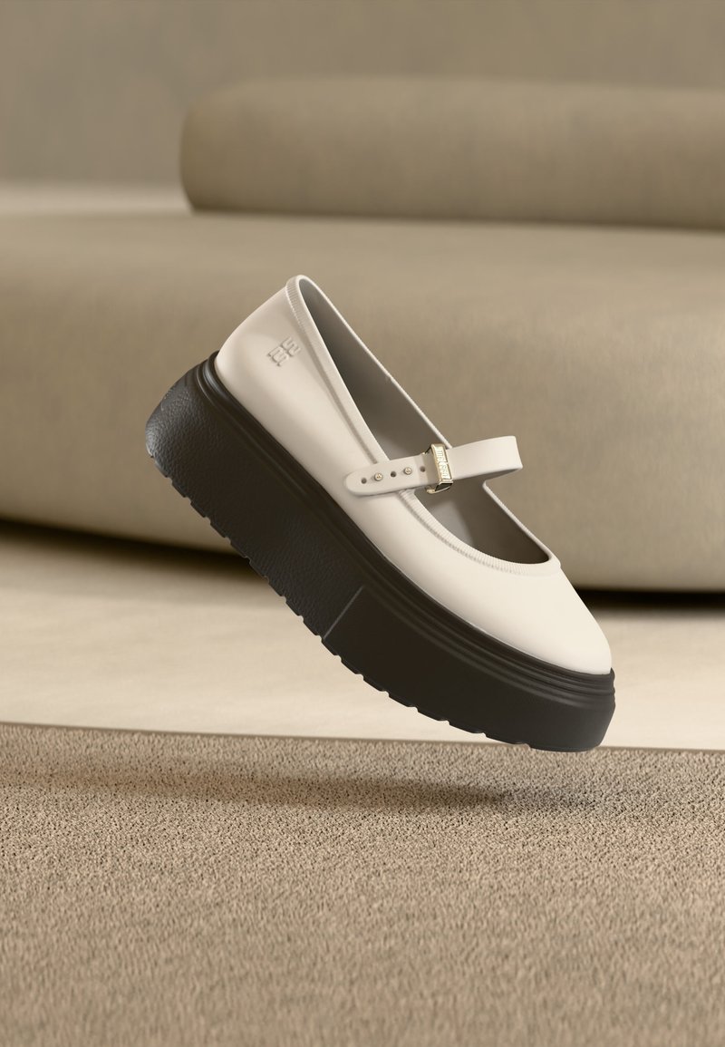 Λευκό παπούτσι Mary Jane με παχύ μαύρο πάτο πλατφόρμας, λουρί με πόρπη, με φόντο μπεζ καναπέ και χαλί.