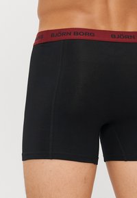 Schwarze Boxer Shorts mit einem bordeauxrotfarbenen elastischen Bund, der den Markennamen "BJÖRN BORG" in Schwarz trägt. Glattes Material mit einer figurbetonten Passform.