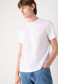 Hvit bomull T-skjorte med rund hals, korte ermer, og rødt "Calvin Klein"-logo på venstre bryst. Kombinert med blå denimjeans.