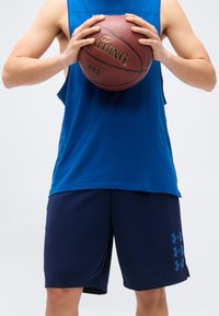 Άτομο με μπλε αμάνικο πουκάμισο και ναυτικό σορτς κρατώντας μια μπάλα μπάσκετ Spalding στο ύψος του στήθους, μπροστά σε απλό φόντο.