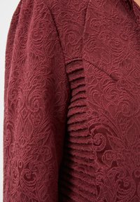 Tejido burdeos texturizado con intrincados patrones florales y pliegues horizontales a lo largo de la manga para añadir detalle y profundidad.