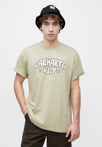 Ανοιχτοπράσινο βαμβακερό t-shirt με ρεβέρ μανίκια, με ένα μεγάλο μαύρο γραφικό κείμενο "Carhartt WIP 1989." Το μοντέλο φοράει μαύρο καπέλο τύπου κάπα.