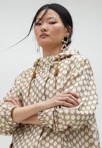 Beżowa i kremowa kurtka z kapturem w kratę z markowym wzorem, z elementami w złotym kolorze i sznurkiem do regulacji. Teksturowana tkanina z elastycznymi mankietami.