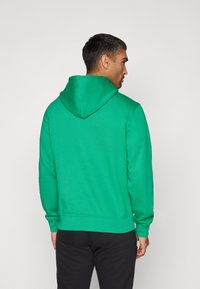 Felpa verde con cappuccio realizzata in tessuto morbido, con tasca a marsupio, polsini a coste e una vestibilità rilassata. Indossata con pantaloni neri.