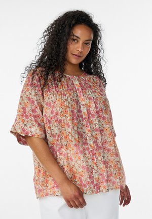 Kvinde med langt krøllet hår iført en løs blomsterprint-bluse i pink, orange og beige nuancer og hvide bukser, stående mod en hvid baggrund.
